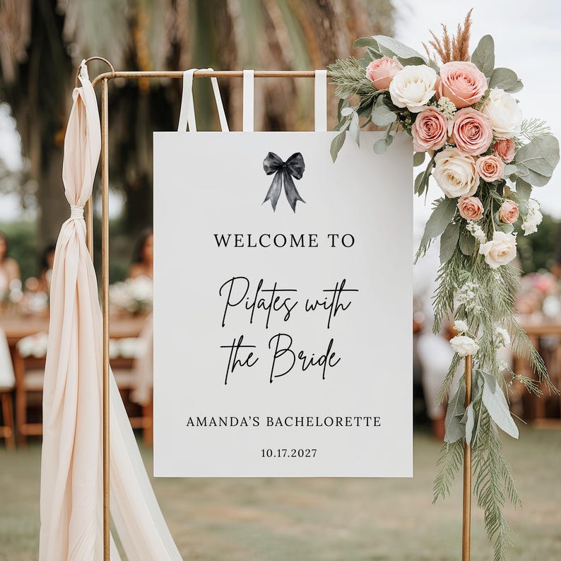May include: A white sign with the text "WELCOME TO Pilates with the Bride" and "AMANDA'S BACHELORETTE 10.17.2027" is displayed on a gold stand. The sign is decorated with a black bow and floral arrangements.