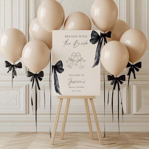 Puede incluir: Un letrero de despedida de soltera con el texto "Brunch with the Bride" y "Welcome to Jessica's Bridal Shower 06.27.2027". El letrero está sobre un caballete de madera, rodeado de globos beige y lazos negros.