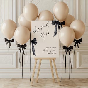 Può includere: Un cartello beige con calligrafia nera che recita "she said yes!" e "Let's Celebrate! Alexis & Brian April 22, 20XX". Il cartello è appoggiato su un cavalletto di legno. Sullo sfondo ci sono palloncini beige con nastri neri legati ad essi.