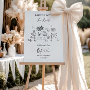 Puede incluir: Un letrero blanco sobre un caballete de madera con el texto "Brunch with the Bride" y "Welcome to Olivia's Bridal Shower 06.27.2027". El letrero presenta dibujos lineales negros de champán, flores, pasteles y una cámara. Un gran lazo color crema adorna el lateral.