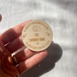 May include: A wooden Parent Decision Coin with engraved text and a baby illustration. The coin reads "PARENT DECISION COIN", "IT'S DADDY'S TURN", and "FLIP FOR YOUR FATE". The coin is held in a hand.