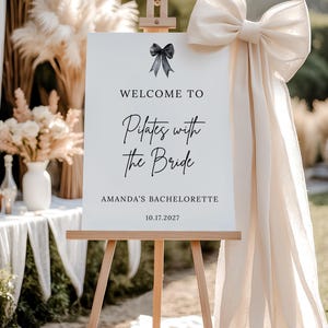 May include: A white sign on a wooden easel with the text "Welcome to Pilates with the Bride" and "Amanda's Bachelorette" with the date 10.17.2027. A large cream-colored bow adorns the right side. Decorative pampas grass and flowers are in the background.