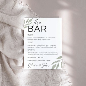 May include: A printable wedding bar sign with a green leafy design. The sign lists the drinks available at the bar, including beer, wine, spirits, and non-alcoholic options. The sign is titled "The Bar" and is signed "Olivia & John".