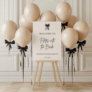 May include: A sign on a wooden easel reads "Welcome to Pilates with the Bride, Amanda's Bachelorette, 10.17.2027." The sign is decorated with a black bow. Beige balloons with black ribbons are arranged around the sign.