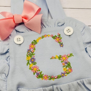 May include: Light blue baby romper with a pink bow and floral embroidered letter "G". The romper has ruffled edges and white buttons. The floral embroidery includes yellow, pink, and purple flowers.