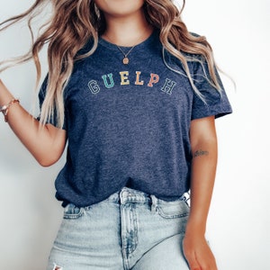 May include: A navy blue t-shirt with the word "GUELPH" printed in rainbow colours across the chest. The shirt is being worn by a person with long blonde hair and blue jeans.