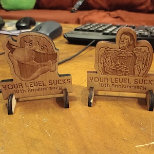 Puede incluir: Dos figuras de madera cortadas con láser con el texto "YOUR LEUEL SUCKS 10th Anniversary". Una presenta un personaje de dibujos animados, la otra una persona. Cada una tiene una pequeña base con ruedas. La madera es de color marrón claro.