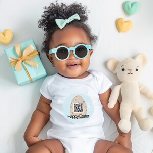 Puede incluir: Un bebé con gafas de sol azul claro y un body blanco con un gráfico de "Felices Pascuas". El gráfico incluye un código QR y un diseño de huevo. Un lazo azul claro, una caja de regalo y un peluche también están en la imagen.