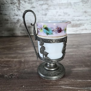 Può includere: Una piccola lattiera in porcellana inserita in una montatura in metallo color peltro con un manico alto. Il bordo superiore della porcellana bianca presenta una fascia dipinta con foglie verdi e bacche viola su sfondo color lavanda.