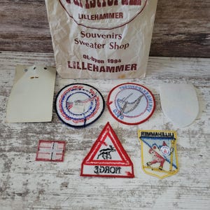 Op de afbeelding: Een verzameling geborduurde patches, waaronder een ronde patch met blauwe en rode randen, een ronde patch met een Vikingschip, een vierkante patch met de Noorse vlag, een driehoekige patch met een eland en de tekst NORGE.