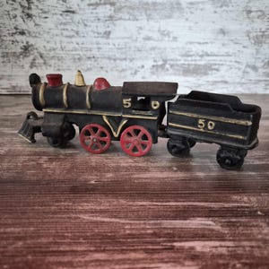 Puede incluir: Una locomotora de vapor de juguete de hierro fundido negro con ruedas rojas y detalles en color dorado. La máquina lleva el número 5 en el lateral, mientras que el ténder de carbón adjunto muestra el número 50. Tiene una chimenea roja y una campana dorada.