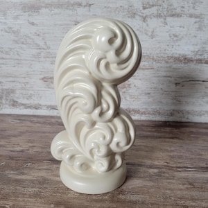 May include: A cream-colored ceramic sculpture featuring a vertical, swirling wave-like design with multiple rounded, embossed scrolls and curves. The object has a smooth, glossy finish and stands on a circular base, showcasing a continuous, fluid form.