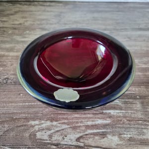 Puede incluir: Un cuenco de cristal circular con un centro rojo rubí profundo y translúcido que se transforma en un borde oscuro, casi negro. El cuenco tiene una superficie lisa y pulida e incluye una pequeña pegatina de color claro con borde festoneado en el borde exterior.