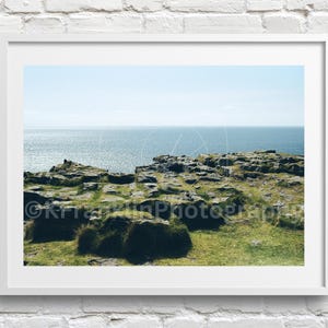 May include: A framed landscape photograph depicting a coastal view. The image shows a rocky cliff with green grass and vegetation, looking out over a vast, shimmering sea under a bright, clear sky. The photograph is presented in a white frame.