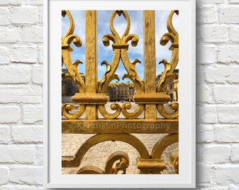 May include: A close-up of a gold-colored metal gate with intricate designs. The gate is open, revealing a view of a building with a courtyard in the background.