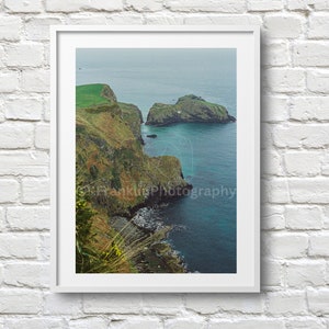 May include: A photograph of a rocky coastline with green grass and a blue ocean. The photo was taken from a high vantage point, looking down at the coastline. The water is calm and the sky is cloudy.