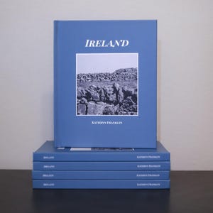 Peut inclure: Plusieurs livres bleus empilés, intitulés "IRELAND". La couverture présente une image en niveaux de gris d'un mur en pierre. Le nom de l'auteur, Kathryn Franklin, est imprimé en dessous. Les livres sont empilés.
