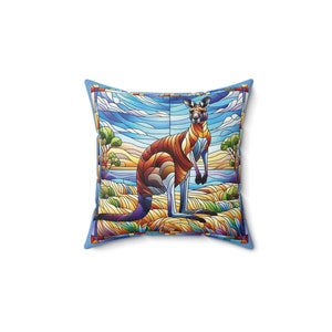 Può includere: Cuscino quadrato con un design a forma di canguro in stile vetrata. Il canguro è raffigurato in tonalità di marrone, arancione e rosso, su uno sfondo di cielo blu, acqua ed erba giallo-verde. Il cuscino ha un bordo azzurro.