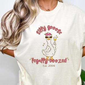 Op de afbeelding: Crèmekleurig T-shirt met een cartoon gans met een zonnebril en een bloemenkrans, met een wijnglas. De tekst luidt "silly goose" erboven en "legally boozed" eronder, met "Est. 2004".