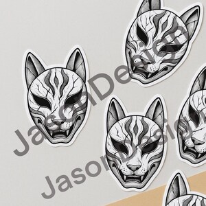May include: A collection of white stickers featuring a stylized, black and white Kitsune mask design. The masks have a fox-like appearance with intricate line details, including the eyes, nose, and mouth. The stickers are arranged on a light gray surface.