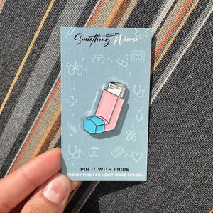 May include: A pink and blue enamel pin shaped like an inhaler with a white label that says "RELEASING AIR". The pin is on a blue card with white text that says "Something Nurse" and "PIN IT WITH PRIDE TRENDY PINS FOR HEALTHCARE HEROES".