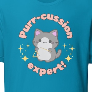 Può includere: Una maglietta blu turchese con un'immagine di un gatto dei cartoni animati che suona un triangolo. Il testo "Purr-cussion expert!" è scritto in un font rosa e bianco intorno al gatto.