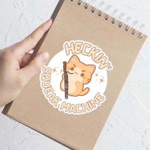 May include: A sticker featuring a cartoon orange cat holding a bassoon. The cat is inside a white circle with the words "Heckin' Squeak Machine" in a playful font. The sticker is on a brown paper background.