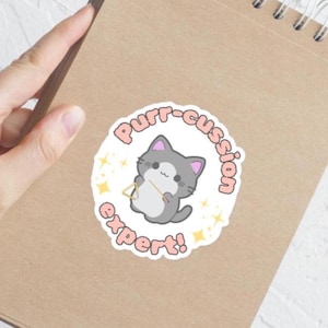 Puede incluir: Una pegatina redonda con un gato gris de dibujos animados tocando un triángulo. La pegatina tiene el texto "Purr-cussion expert!" en rosa, con estrellas. La pegatina está sobre un cuaderno marrón. El diseño es divertido y musical.