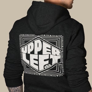 May include: Dark gray hoodie with a white graphic on the back that reads "UPPER LEFT USA" inside a geometric design. The hoodie has a hood and a front zipper. The sleeves have ribbed cuffs.