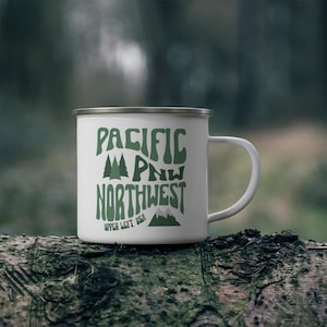 May include: White enamel mug with a silver rim and a large white handle. The mug features the words "PACIFIC PNW NORTHWEST" in green, with tree and mountain graphics. The text "UPPER LEFT USA" is below the word "NORTHWEST".