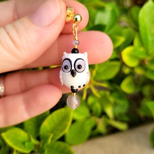 May include: A gold-colored stud earring with a dangling white owl charm. The owl has large black eyes and small black dots. A small gray bead and a larger, round, light purple bead hang below the owl. The earring is held against a background of green leaves.