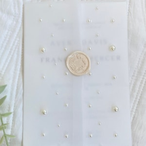 May include: A white wedding invitation with a wax seal and pearl embellishments. The invitation reads "[Name] & [Name]  [Date] [Time] [Location]"