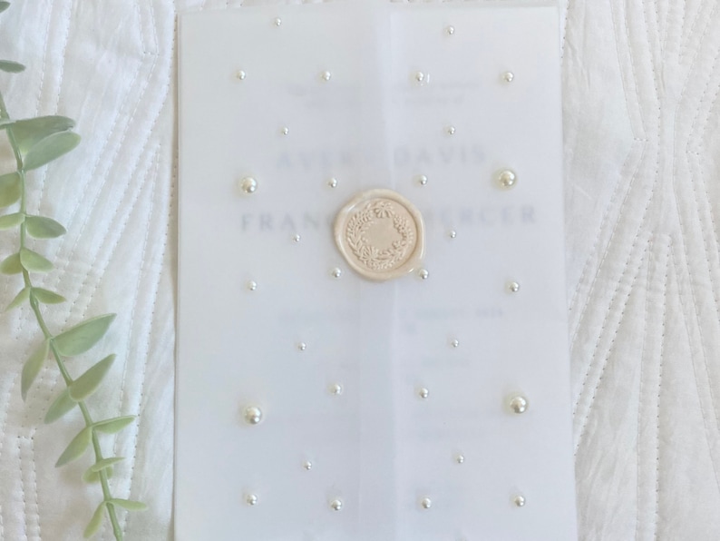 May include: A white wedding invitation with a wax seal and pearl embellishments. The invitation reads "[Name] & [Name]  [Date] [Time] [Location]"