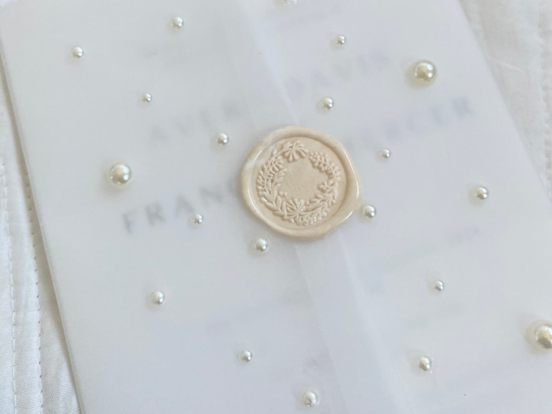 May include: A white envelope with a wax seal and pearl embellishments. The wax seal features a floral design.