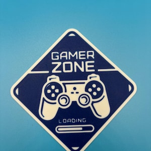 Może przedstawiać: Niebiesko-biała naklejka z napisem "GAMER ZONE" i "LOADING". Naklejka przedstawia białą grafikę kontrolera pośrodku. Naklejka ma kształt rombu i białą obwódkę.