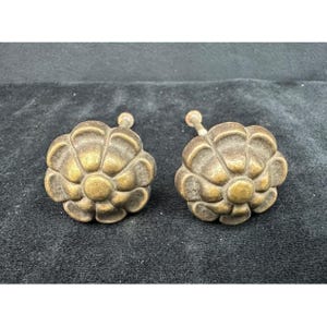 May include: Two antique brass flower-shaped cabinet knobs. Each knob features a detailed floral design with a central button and a stem for attachment. The knobs have a warm, aged patina, suggesting vintage craftsmanship.