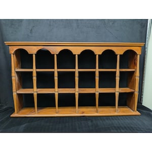 May include: A wooden wall shelf with a warm, medium-brown finish. The shelf features multiple small compartments, supported by vertical spindles and horizontal shelves. The top has an arched design, adding a decorative element to the storage unit.