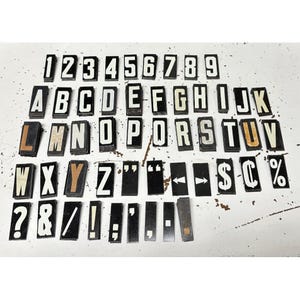 May include: A collection of vintage black and white letterpress printing blocks, including numbers, letters, and punctuation marks. The blocks are arranged on a white surface, showcasing their bold, sans-serif fonts. The set includes a variety of symbols.