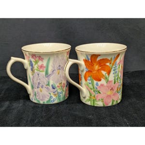 May include: Two ceramic mugs with a cream-colored base and gold-trimmed rims. The left mug features purple iris flowers and green foliage. The right mug displays vibrant orange and pink lily flowers with green stems and blue accents.