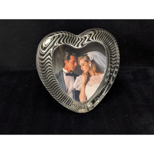 May include: A heart-shaped glass picture frame featuring a clear, wavy, rippled texture along the border. The frame contains a photograph of a couple, with one person wearing a suit and the other wearing a white wedding dress and veil.