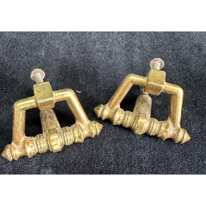 May include: Two antique brass drawer pulls. Each pull has a rectangular mounting block with a screw, a curved handle, and a decorative base. The pulls are tarnished with age and wear.