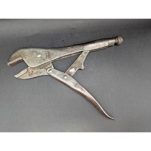 May include: A vintage metal locking pliers tool with a weathered finish. The tool features a serrated jaw, a release lever, and an adjustment screw at the end of the handle. The text Vise-Grip is embossed on the side of the metal body.