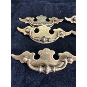 May include: A collection of antique brass drawer pulls. Each pull features a decorative, curved design with a central handle and ornate detailing. The brass has a warm, aged patina, suggesting vintage craftsmanship.