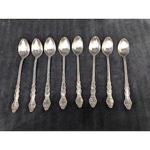 May include: A set of eight silver-toned metal spoons arranged in a horizontal row. Each spoon features a long, slender handle with a decorative, embossed pattern near the base and a smooth, oval-shaped bowl at the top for serving or eating.