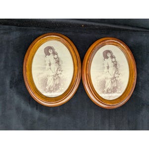 May include: A pair of matching oval wooden picture frames with a warm brown finish. Each frame contains a sepia-toned print of a person wearing a wide-brimmed hat and a long dress, holding a bouquet of flowers while standing in a field.