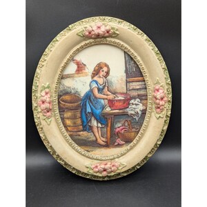 May include: Oval-shaped wall art featuring a painting of a young woman washing clothes. The artwork is set within a cream-colored frame adorned with pink floral accents. The painting depicts a rustic scene with barrels and a basket.
