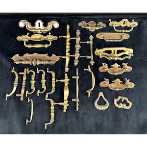 May include: A collection of antique brass drawer pulls and handles in various styles. The hardware includes ornate pulls with keyholes, curved handles, and decorative backplates. The pieces are arranged on a black surface.
