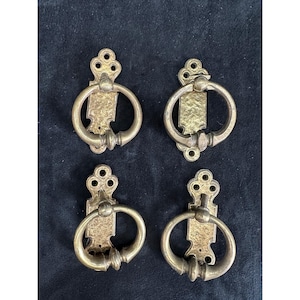 May include: Four antique brass drawer pulls. Each pull features a decorative backplate with a ring handle. The brass has a warm, aged patina. The pulls are likely for a cabinet or drawer.