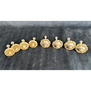 May include: A collection of eight antique brass drawer pulls. Each pull features a decorative, floral-inspired design with a small, round knob. The pulls are arranged in a row against a dark background, showcasing their aged, golden color.
