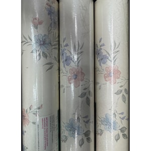 May include: Three rolls of floral wallpaper. The wallpaper features a repeating pattern of pink and blue flowers with green leaves on a cream background. The rolls are tightly wound and wrapped in clear plastic. The text on the label reads "MADE IN CANADA BY FABRIQUE AU C".
